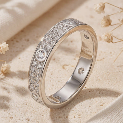 Silver ring standing on a beige textured surface, showing its pavé front, round bezel-set stones, and smooth inner band.