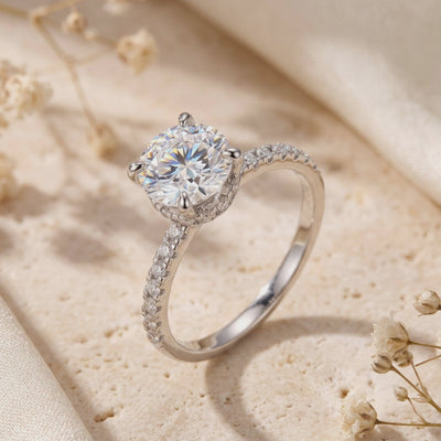 Angled tabletop view of a silver ring with a round clear stone, four-prong basket, and pavé-set shoulders.