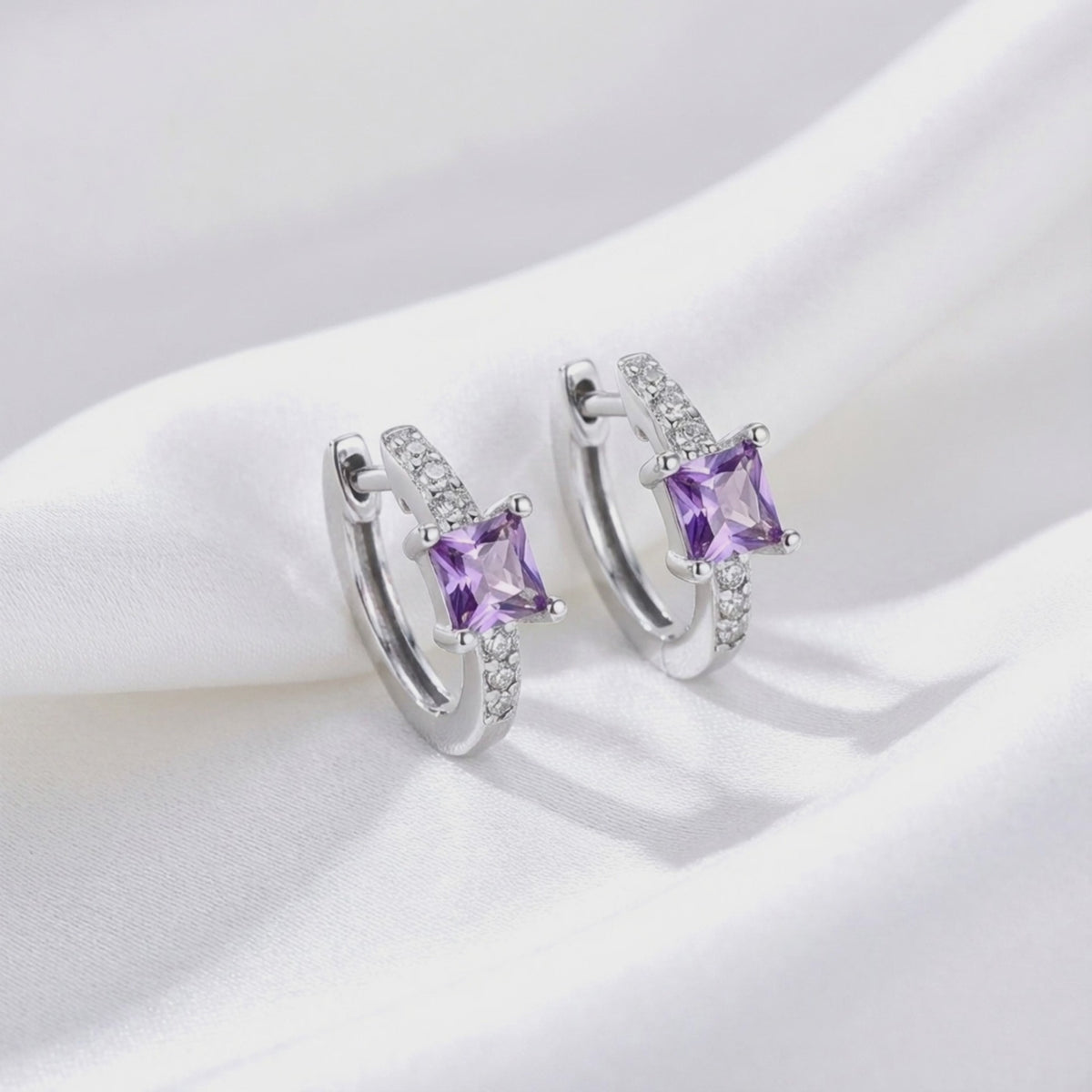 Pair of solid white gold huggie earrings with square purple simulated amethyst center stones and clear accent stones displayed upright on white fabric.