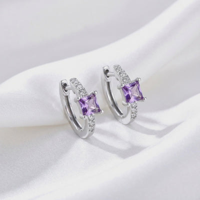 Pair of solid white gold huggie earrings with square purple simulated amethyst center stones and clear accent stones displayed upright on white fabric.
