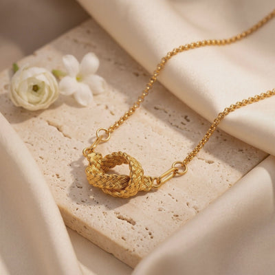 Gold-tone braided knot necklace arranged on a textured stone slab with cream fabric and white flowers.