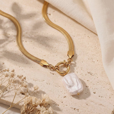 Flat lay of a gold herringbone chain necklace with a white baroque pearl pendant on a textured stone surface.