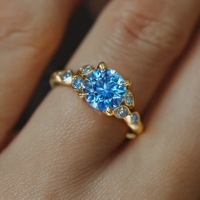 A hand wearing The Katalina solid gold ring with a round blue moissanite center stone and smaller side stones in claw settings.