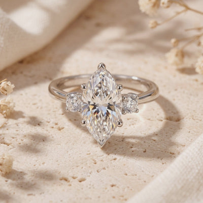Silver three-stone ring with a marquise center stone and round accents, displayed upright on a pale textured surface.