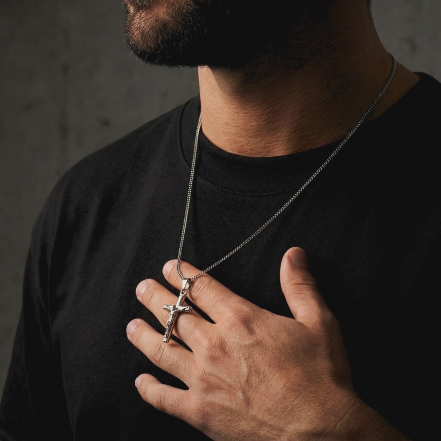 Man in a black shirt holds a silver crucifix pendant on its chain against his chest.