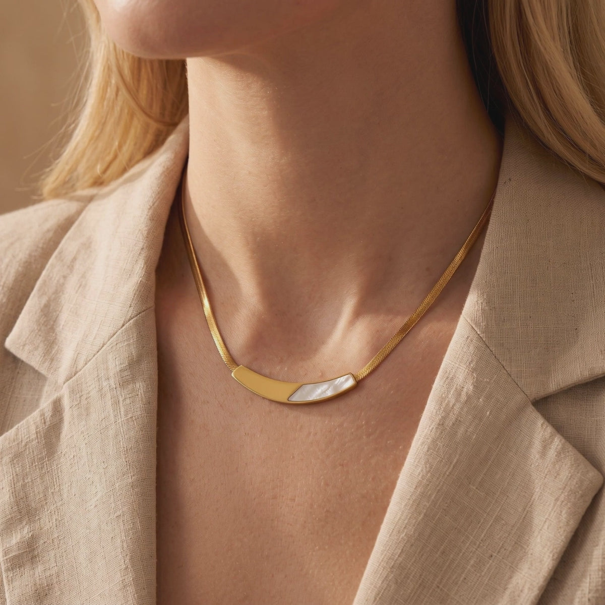 Gold snake chain necklace with a curved mother-of-pearl bar, shown worn on a model in a beige blazer.