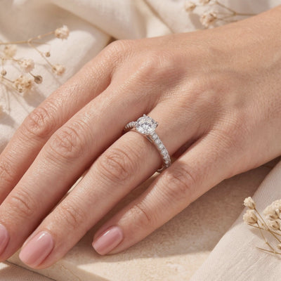 Silver solitaire ring with a round clear center stone and pavé shoulders worn on a hand among dried flowers.