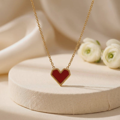 Gold chain necklace with a red heart pendant displayed on a textured beige pedestal beside white flowers.