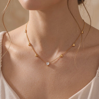 Gold-tone chain necklace with small heart charms and a clear heart pendant worn around a person’s neck.