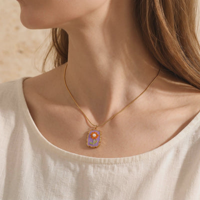 Model wearing Amorina necklace with purple floral pendant, orange flower, green leaves, and simulated pearl center.