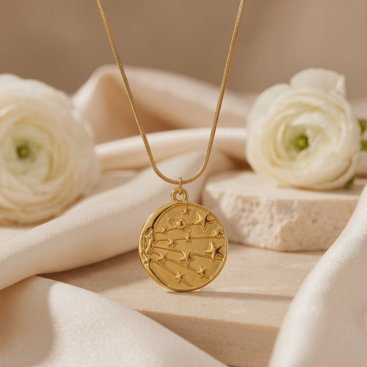 Close-up of a gold-tone celestial medallion necklace displayed on cream fabric beside white flowers.