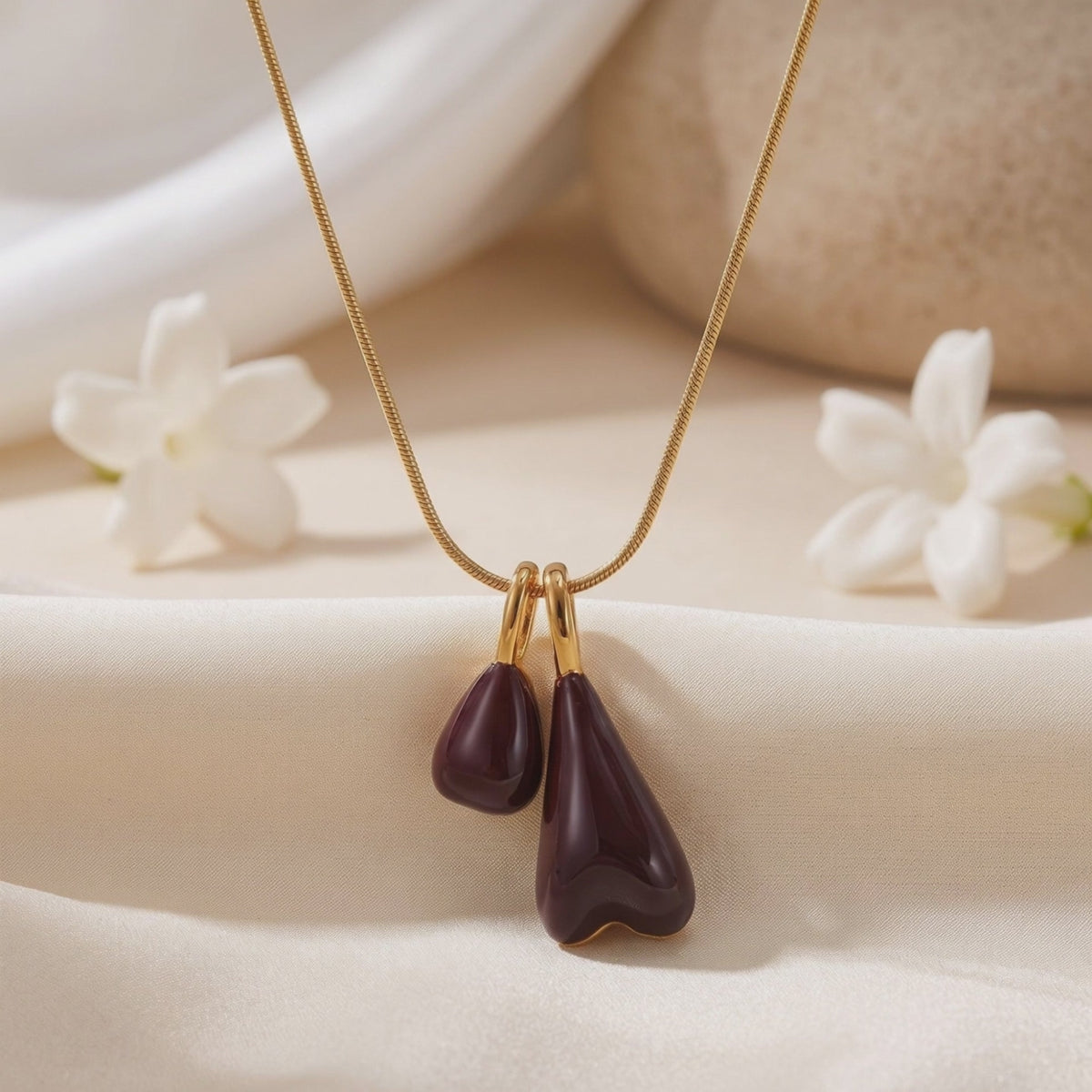Close-up of The Marais gold necklace with two glossy burgundy pendant drops on off-white fabric.