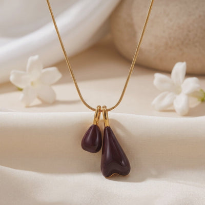 Close-up of The Marais gold necklace with two glossy burgundy pendant drops on off-white fabric.