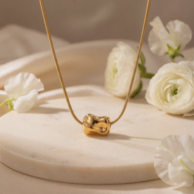 Video preview of a gold snake chain necklace with an organic molten pendant on a round pale pedestal among white flowers.