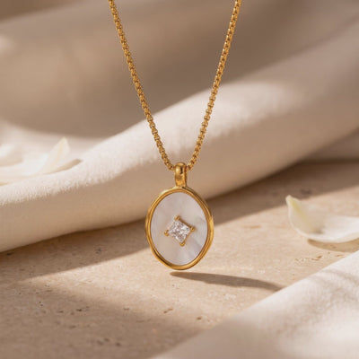 Close-up of a gold-tone necklace with an oval mother-of-pearl pendant and square center stone on a beige surface.