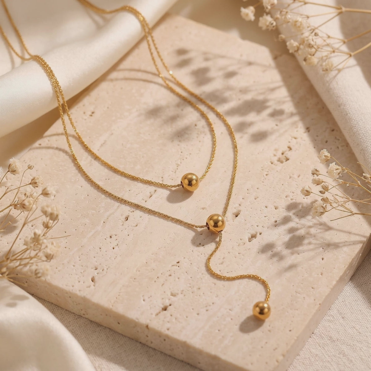 Gold-toned layered lariat necklace with sphere beads arranged on a stone tile beside dried flowers.