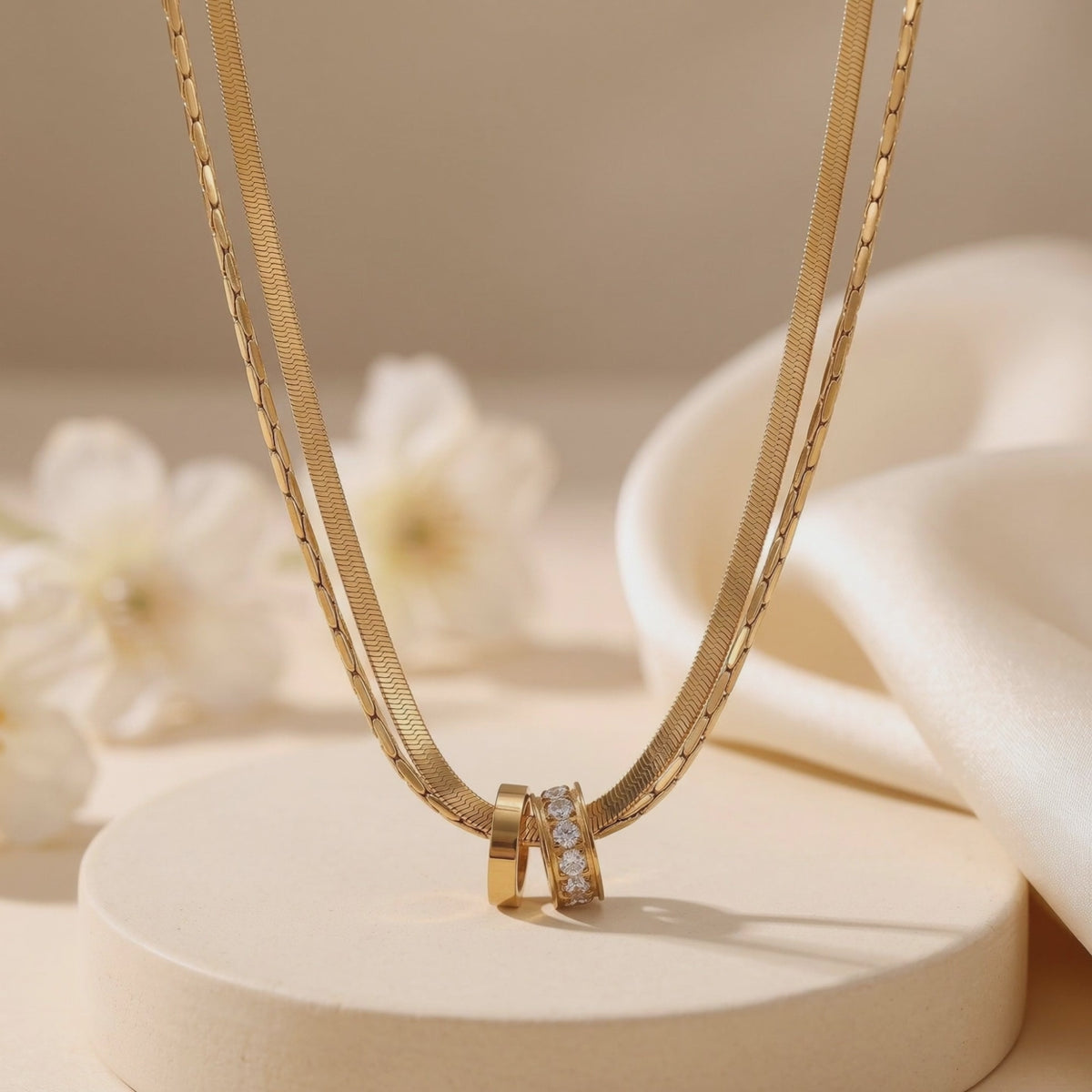 Gold-finish layered necklace with two interlocking ring pendants displayed on a beige pedestal.