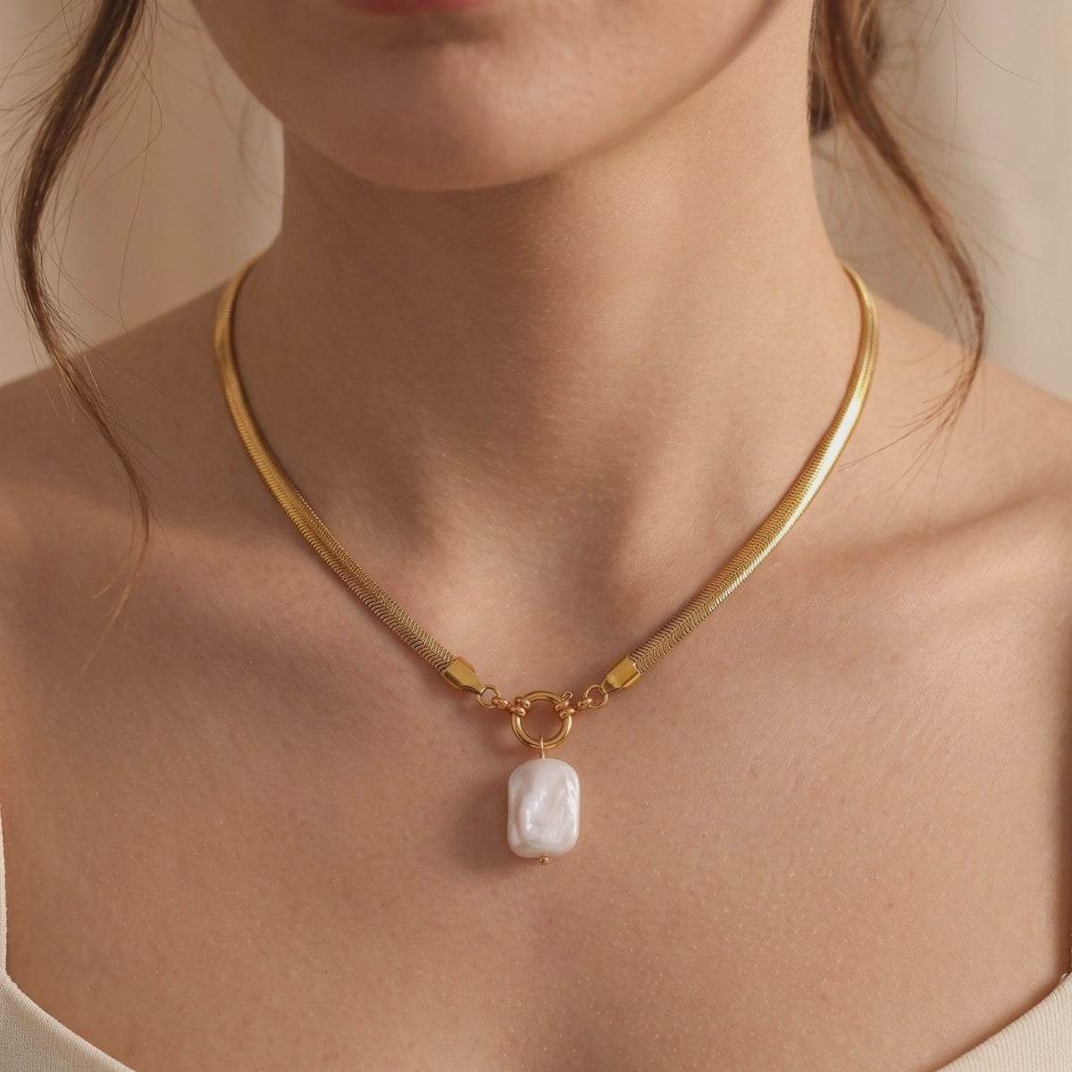 Gold herringbone snake chain necklace with a white baroque pearl pendant, worn around a woman's neck.