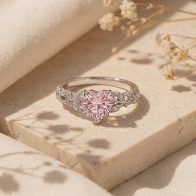 Close-up view of The Rosalique Ring, a solid gold ring with a heart-shaped pink VVS Moissanite centerpiece and floral filigree band, resting on a stone surface.