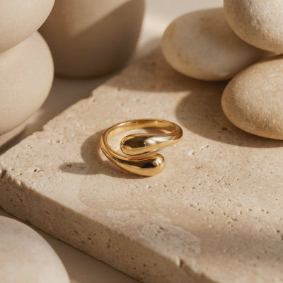 A solid gold open ring with rounded ends resting on a textured beige stone slab among smooth stones.