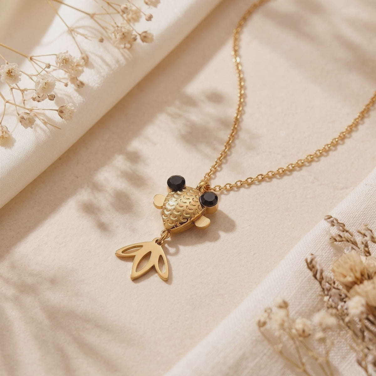 Gold-tone goldfish pendant necklace with black eyes arranged on cream fabric beside dried flowers.