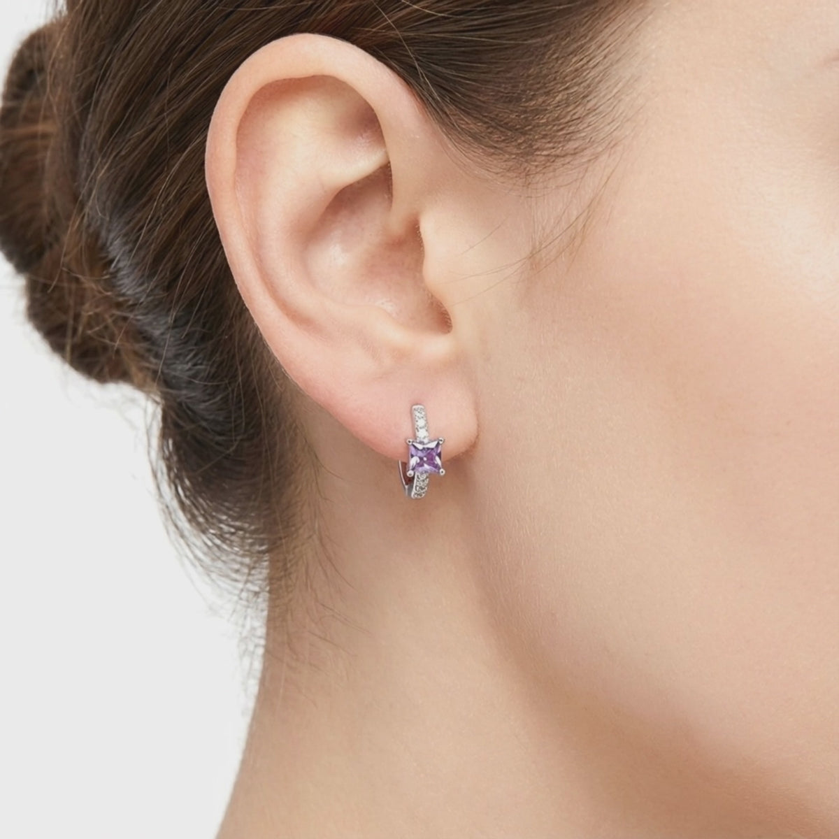 Model wearing a solid white gold huggie earring with a square purple simulated amethyst center stone and clear accent stones on the earlobe.