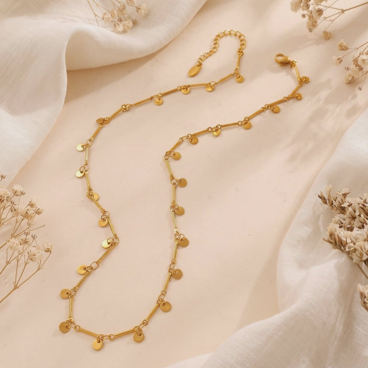 Gold-tone disc charm bar chain necklace arranged on off-white fabric with dried flowers.