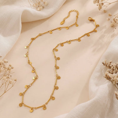 Gold-tone disc charm bar chain necklace arranged on off-white fabric with dried flowers.