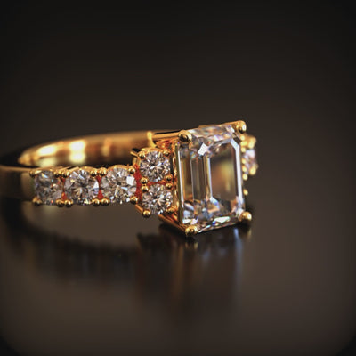 A solid gold ring with an emerald-cut center stone and small round side stones on a dark reflective surface.