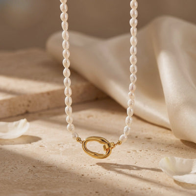 White rice pearl necklace displayed hanging above a warm textured surface, with its gold-tone loop clasp at the bottom.